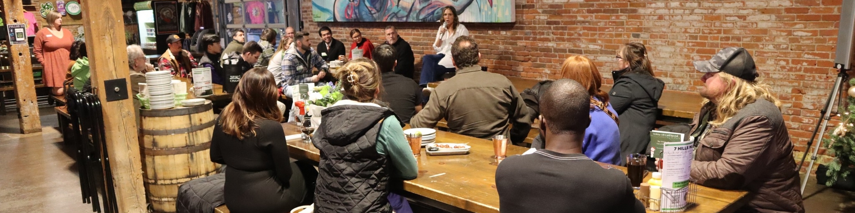 people sitting at a long table inside a brewery listening to a speaker